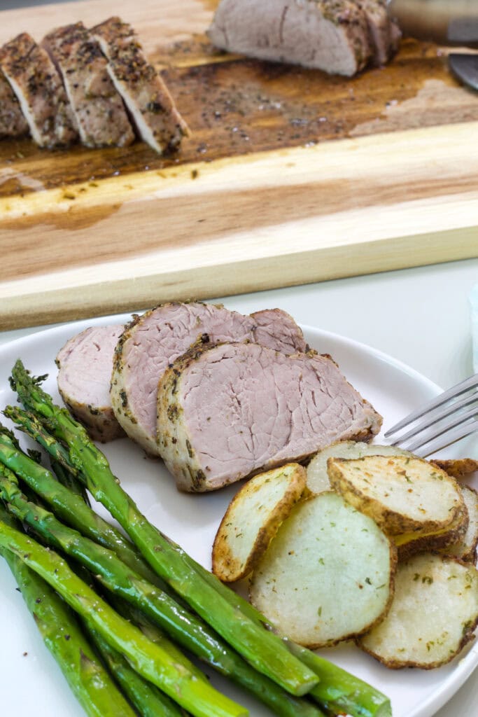 Three slices of pork tenderloin on a plate with air fried potatoes and roasted asparagus spears.