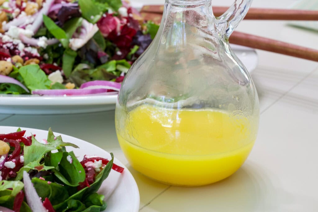 A small dressing bottle with orange vinaigrette in it sitting in near a platter and plate with salad on them.