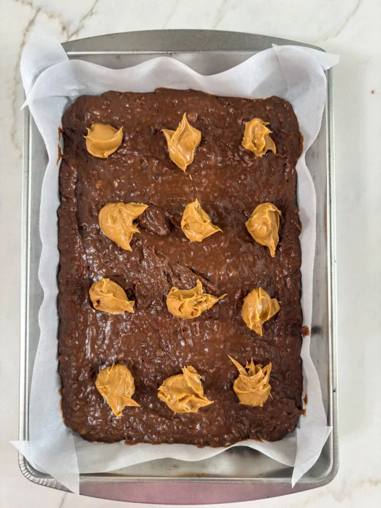 The brownie batter in the prepared baking pan with twelve dollops of peanut butter on top.