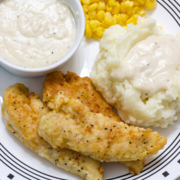 Feature image of three chicken tenders on a plate with a bowl of gravy, mashed potatoes and gravy and corn.