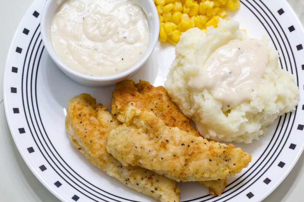 Three Fried Chicken Tenders with White Gravy on a plate with mashed potatoes and corn.