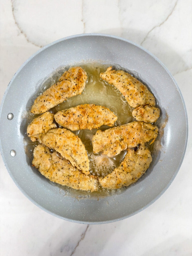 Seven breaded chicken fingers in the skillet after they've cooked on one side and been flipped over.