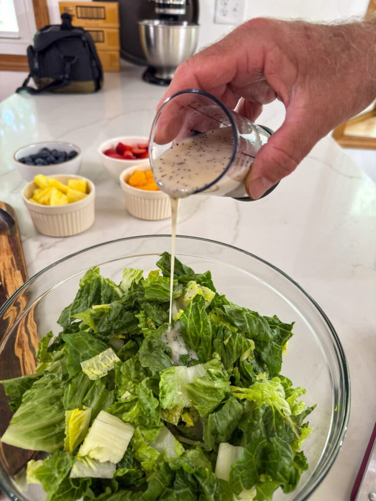 The poppyseed dressing being poured into a glass bowl filled with chopped romaine lettuce.