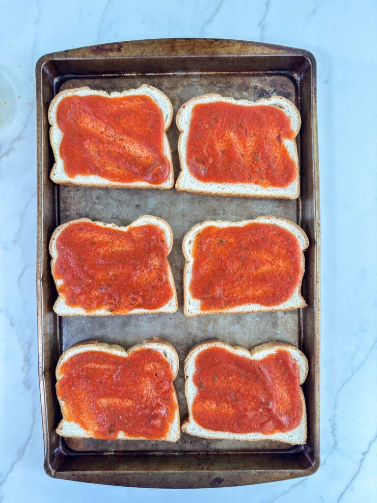 Six slices of bread topped with pizza sauce on a rimmed baking sheet.