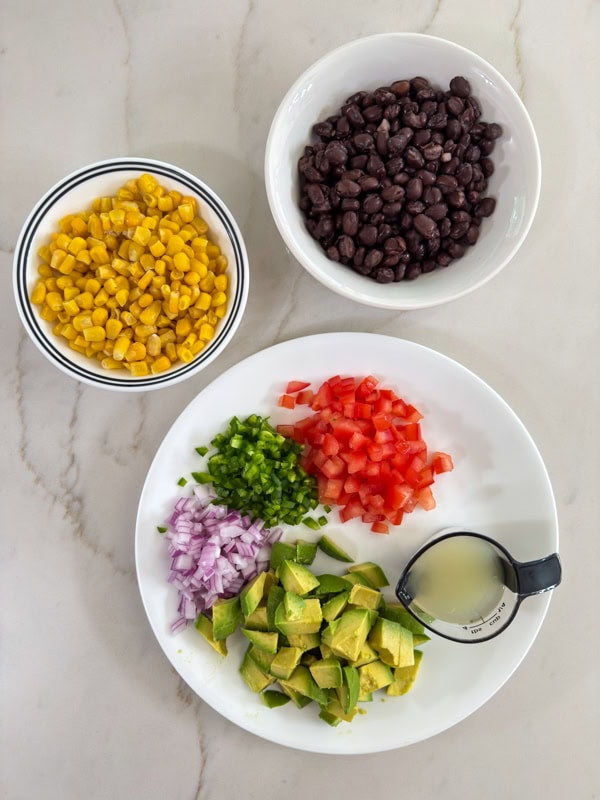 The avocado, onion, tomato and jalapeno chopped on a plate and the corn and black beans in separate bowls.