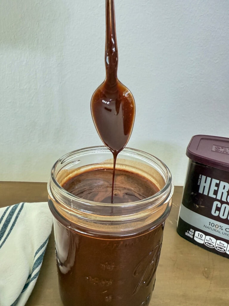 Some chocolate syrup dripping off a spoon into the jar with the rest of the syrup.