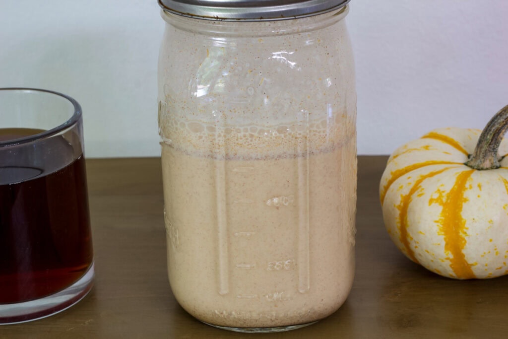 A mason jar with the whole batch of Pumpkin Spice Creamer in it with a cup of coffee on one side and a small pumpkin on the other.