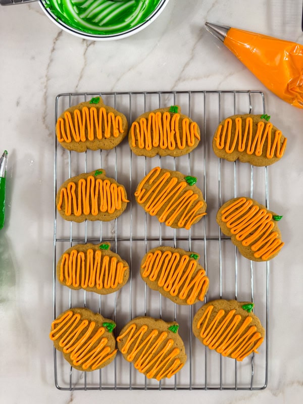 Eleven frosted Pumpkin Spice Cutout Cookies on a black wire cooling rack with piping bags next to it.