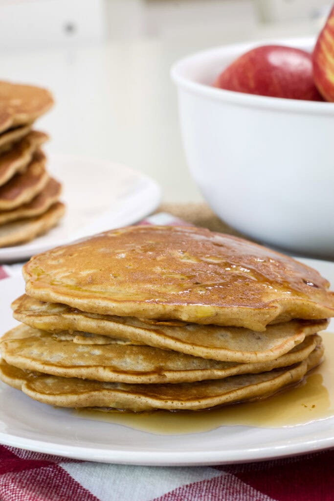 A stack of Apple and Cinnamon Pancakes in the foreground and stack of several more and a bowl of apples in the background.