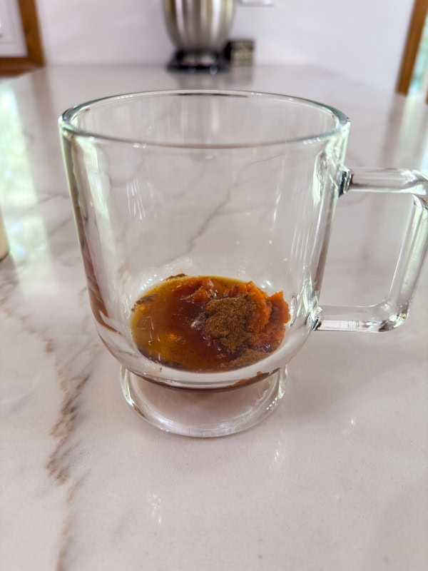 The pumpkin, maple syrup, vanilla and pumpkin pie spice in the bottom of a clear glass coffee mug.
