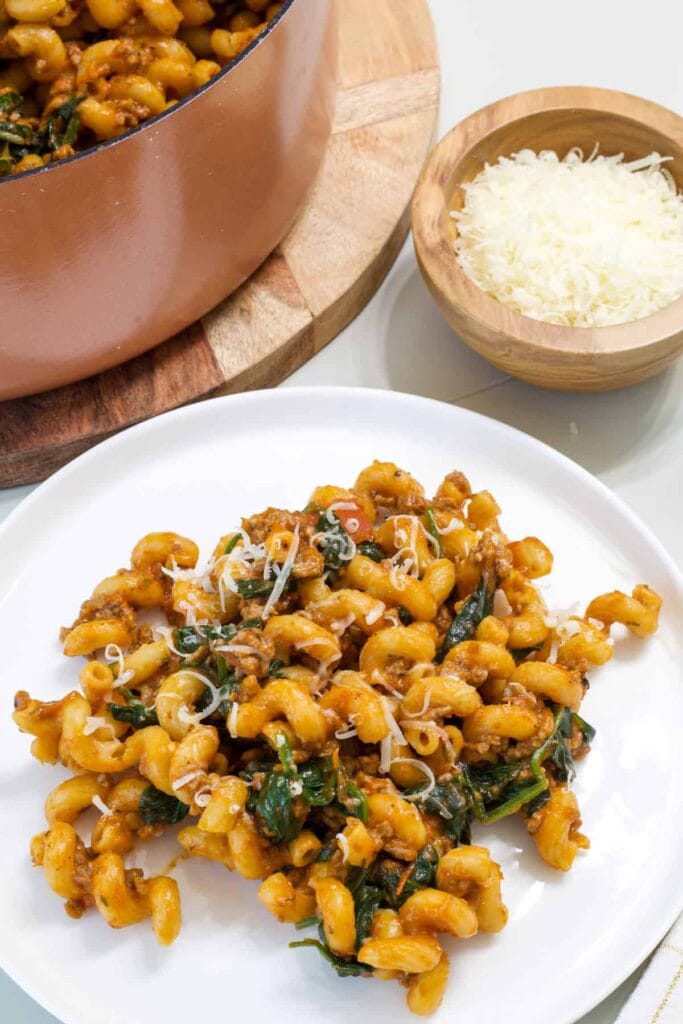 A serving of Ground Beef Spinach Pasta on a plate with a small bowl of parmesan cheese next to it.