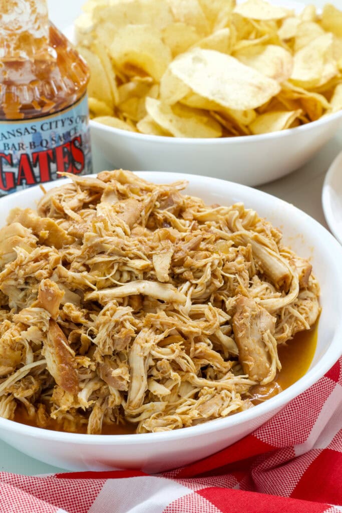 A bowl of Slow Cooker Barbecue Chicken and a bowl of chips and bottle of bbq sauce behind it.