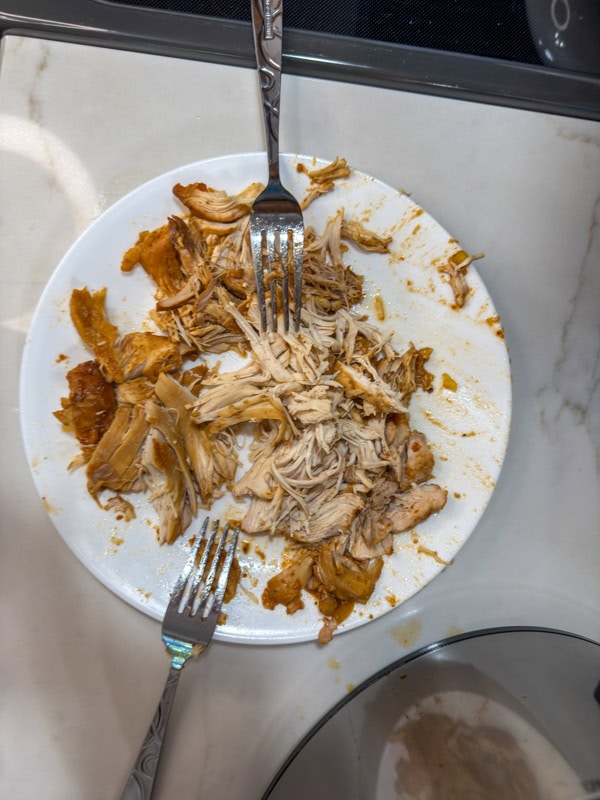 Some of the chicken on a white plate after it's been shredded with two forks.