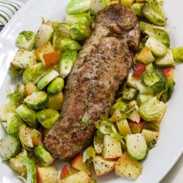 The juicy seasoned uncut pork tenderloin on a platter surrounded by roasted Brussels sprouts and apples.