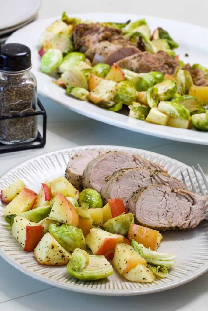 A serving of pork with apples and Brussels sprouts on a plate and the platter with the remaining in the background.