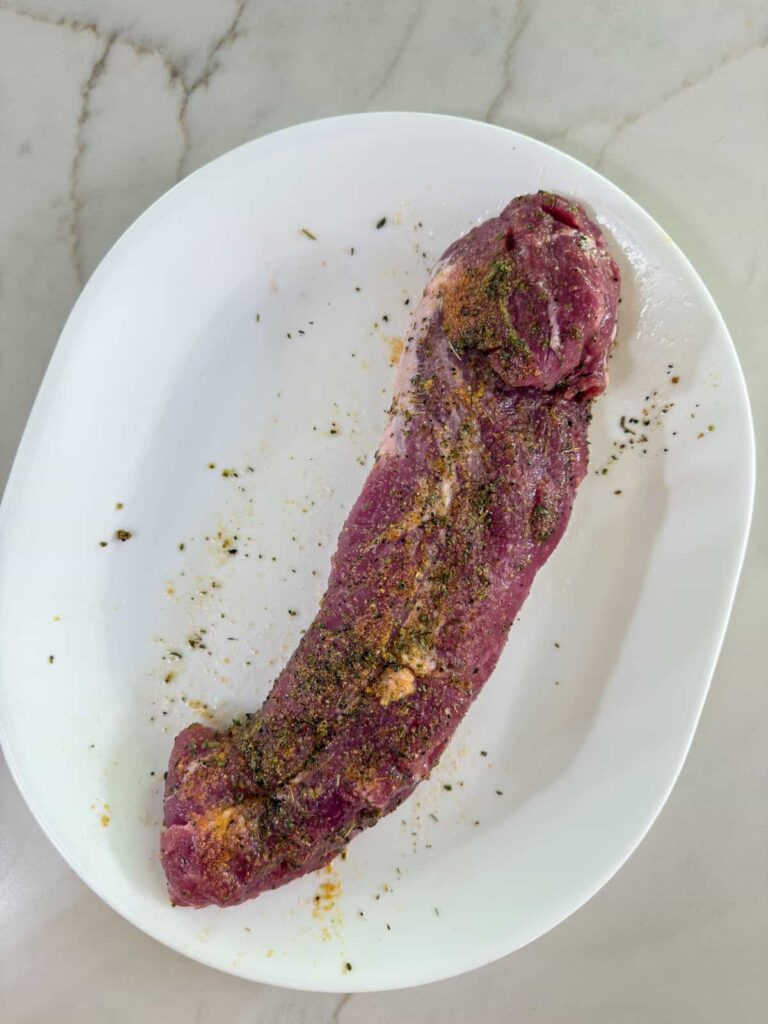 The raw pork tenderloin on a platter after it's been drizzled with olive oil, garlic powder and Italian seasoning.