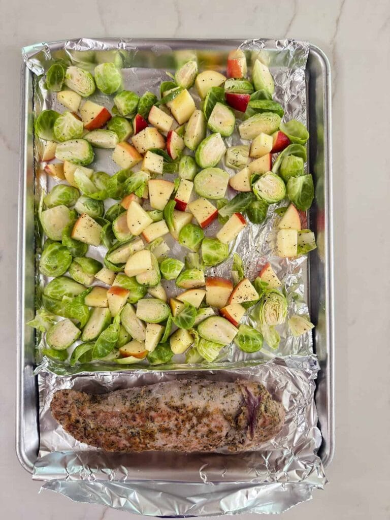 The seasoned pork tenderloin, Brussels sprouts and apples on a foil lined sheet pan ready to go into the oven.