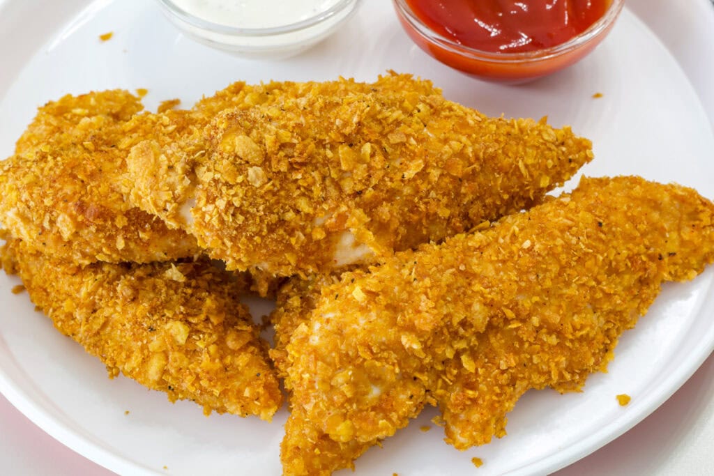 Four Crispy Baked Cornflake Chicken Strips on a plate with two small bowls of dipping sauce.