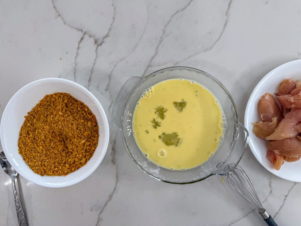 The crushed cornflakes in a bowl, the egg mixture in a shallow dish and the chicken tenders on a plate ready to be coated.