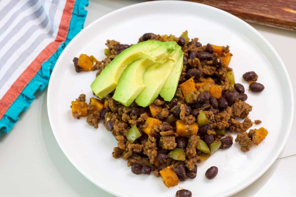 One serving of Ground Beef Sweet Potato Hash on a white plate with a few avocado slices on it.