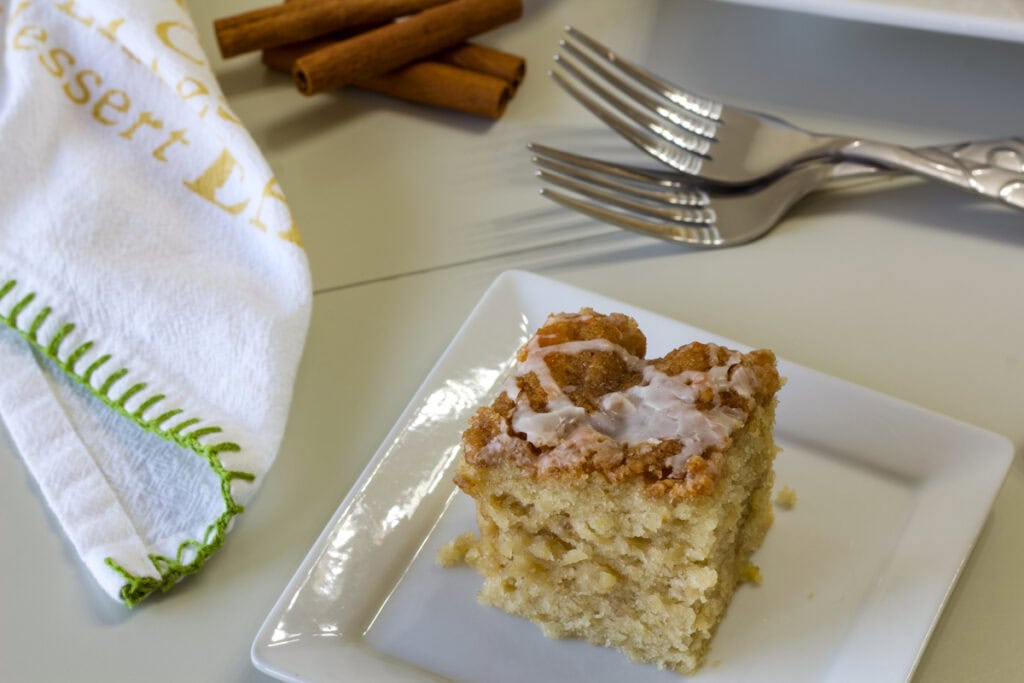 Delicious homemade banana sour cream coffee cake served on a white plate with cinnamon sticks and forks in the background, perfect for breakfast or dessert.