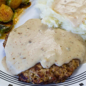 Very close up shot of the Chicken Fried Steak with Cream Gravy over mashed potatoes on a white and black plate.