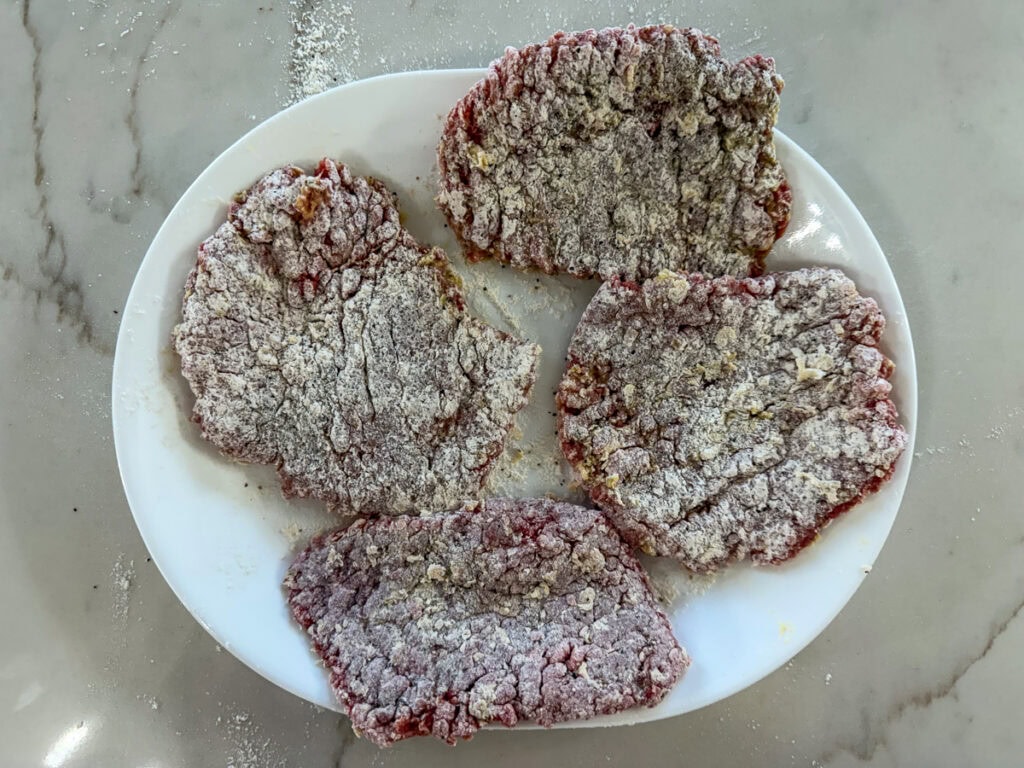 Four raw breaded steaks resting on a white plate.