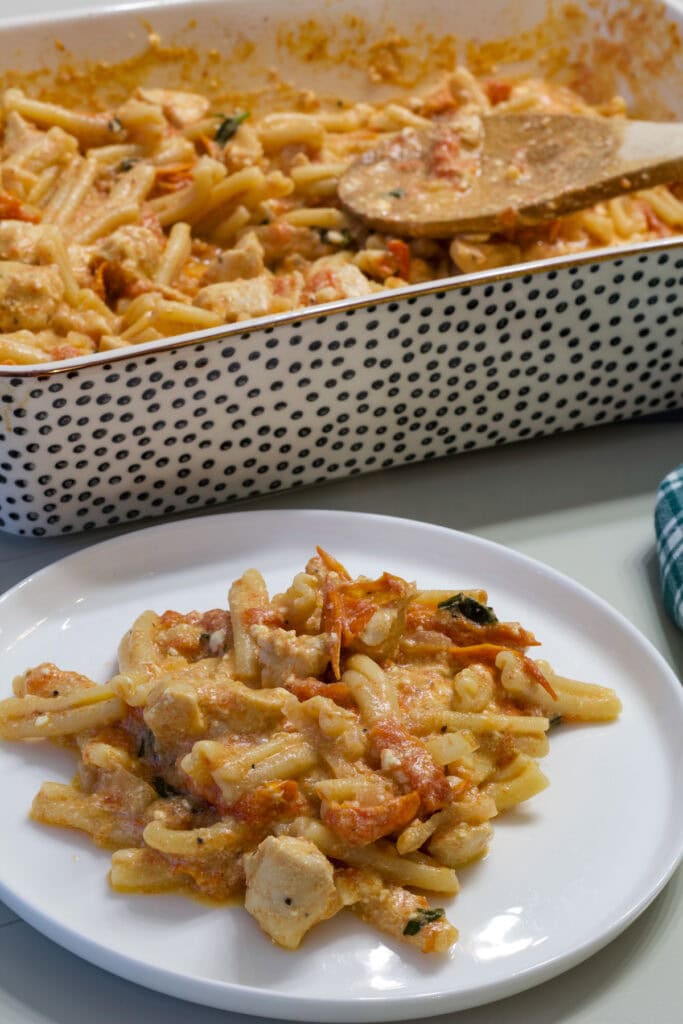 A serving of Easy Baked Feta Pasta with Chicken on a plate in the foreground and the casserole dish with the remaining in the background.