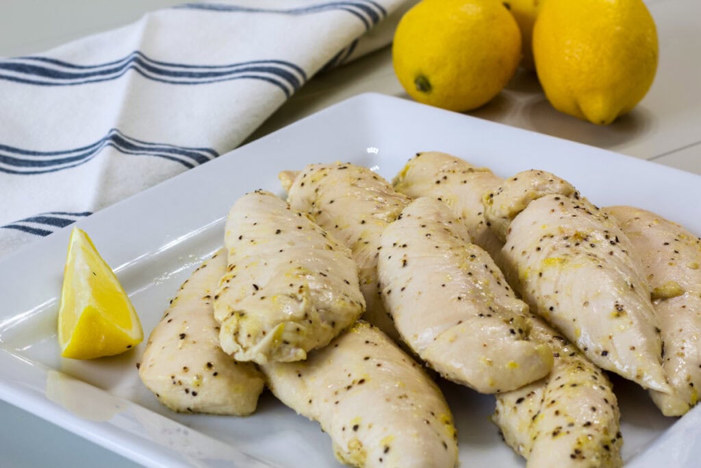 Several Lemon Pepper Chicken Strips on a white plate with a lemon wedge next to them.