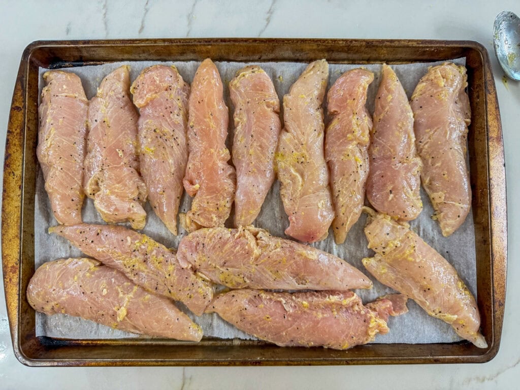 The marinated chicken tenderloins on a parchment lined baking sheet ready to go into the oven.