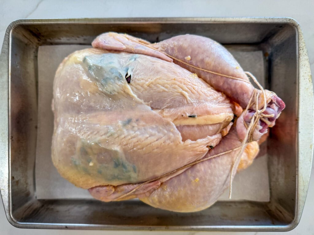 A whole raw chicken prepared for roasting, placed in a metal baking pan, ready for seasoning and cooking.