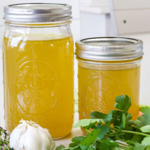 Two jars of homemade chicken stock with a head of garlic and fresh herbs in front of them.