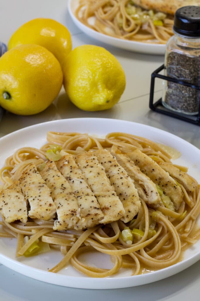 Side shot of a plate with wheat linguine topped with a sliced piece of pan fried chicken.