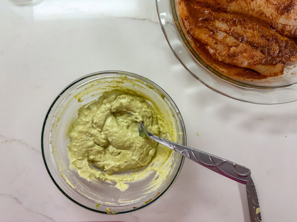 Avocado cream for the simple swai fish taco recipe in a small bowl with the fish marinating in a pie pan nearby.
