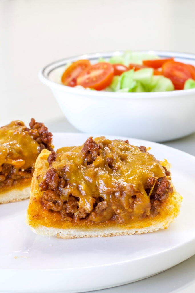 Two pieces of Sloppy Joe Garlic Bread on a plate in the foreground and a bowl of salad in the background.