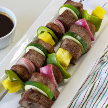 Overhead close up of two baked Teriyaki Beef Skewers on a serving platter with a small bowl of sauce next to it.
