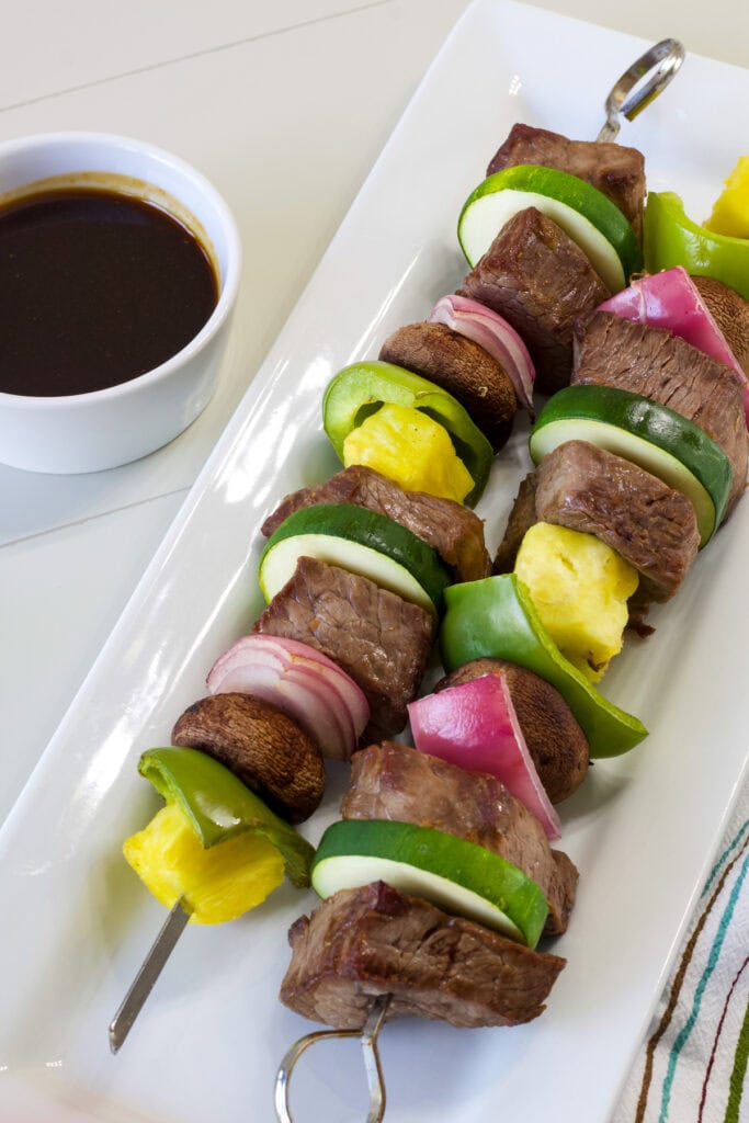 Two Teriyaki Beef Skewers on a white serving plate with a bowl of teriyaki sauce next to it.