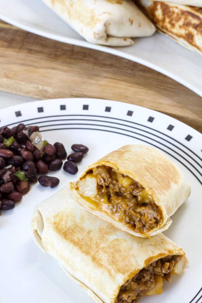 One cut Cheesy Beef and Potato Burritos so the inside is visible on a plate with black beans.
