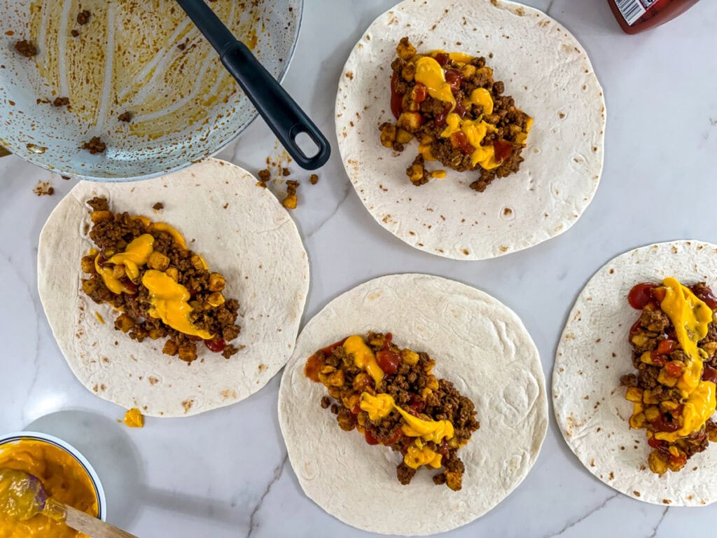 Four flour tortillas filled with cheese sauce, taco sauce and the ground beef potato mixture, ready to be rolled up.