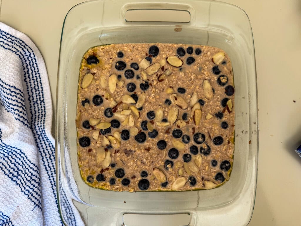 Blueberry baked oatmeal mixture in a glass baking dish topped with fresh blueberries and sliced almonds before baking.