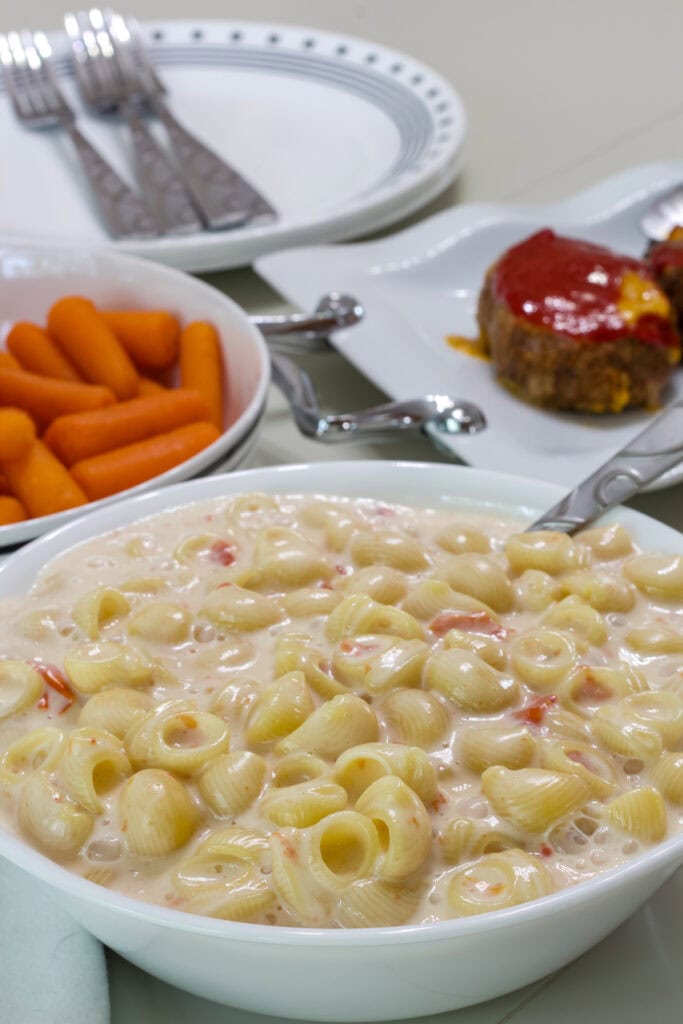 A bowl of Queso Blanco Mac and Cheese and a bowl of carrots and plate of meatloaf in the background.