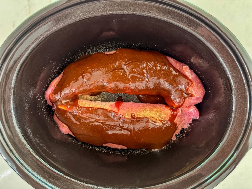 The pork tenderloins with rib rub and barbecue sauce on them in the slow cooker before being cooked.