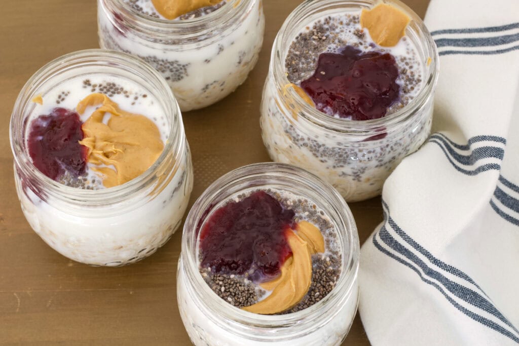 Four small mason jars with all of the ingredients for PB&J Overnight Oats before being stirred.
