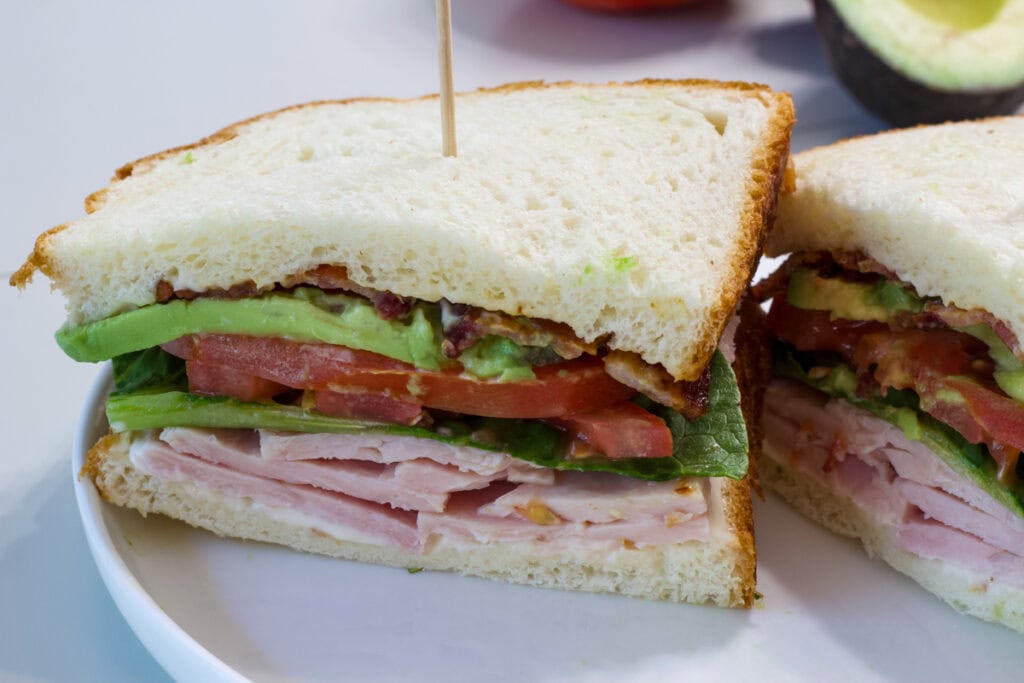 A turkey avocado blt sandwich on a plate cut in half so the layers of ingredients are visible.