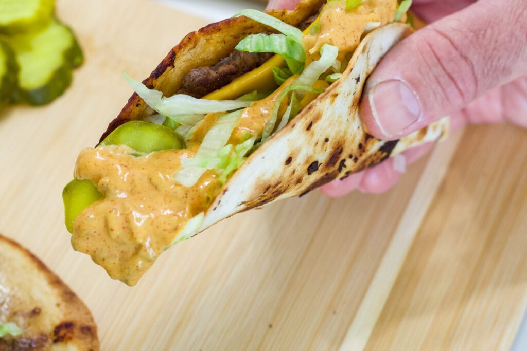Close-up of a hand holding a Big Mac smash taco with melted cheese, lettuce, pickles, and Big Mac sauce.