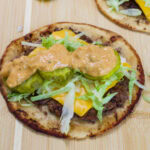 Close-up of a homemade big mac smash taco topped with pickles, shredded lettuce, cheese, and creamy big mac sauce on a wooden cutting board.