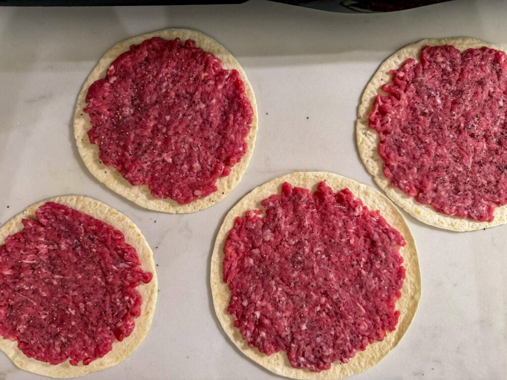 Four flour tortillas topped with raw ground beef seasoned with salt and pepper, ready to be cooked.
