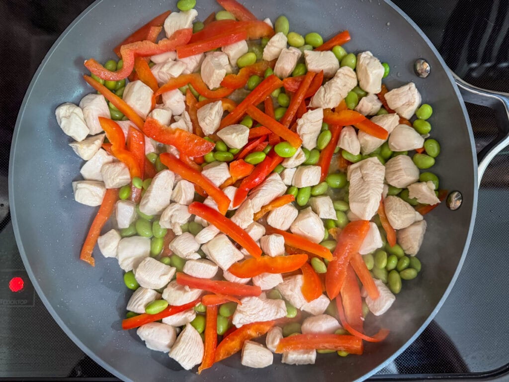 Red bell pepper strips and edamame added to the cooked chicken in a skillet.