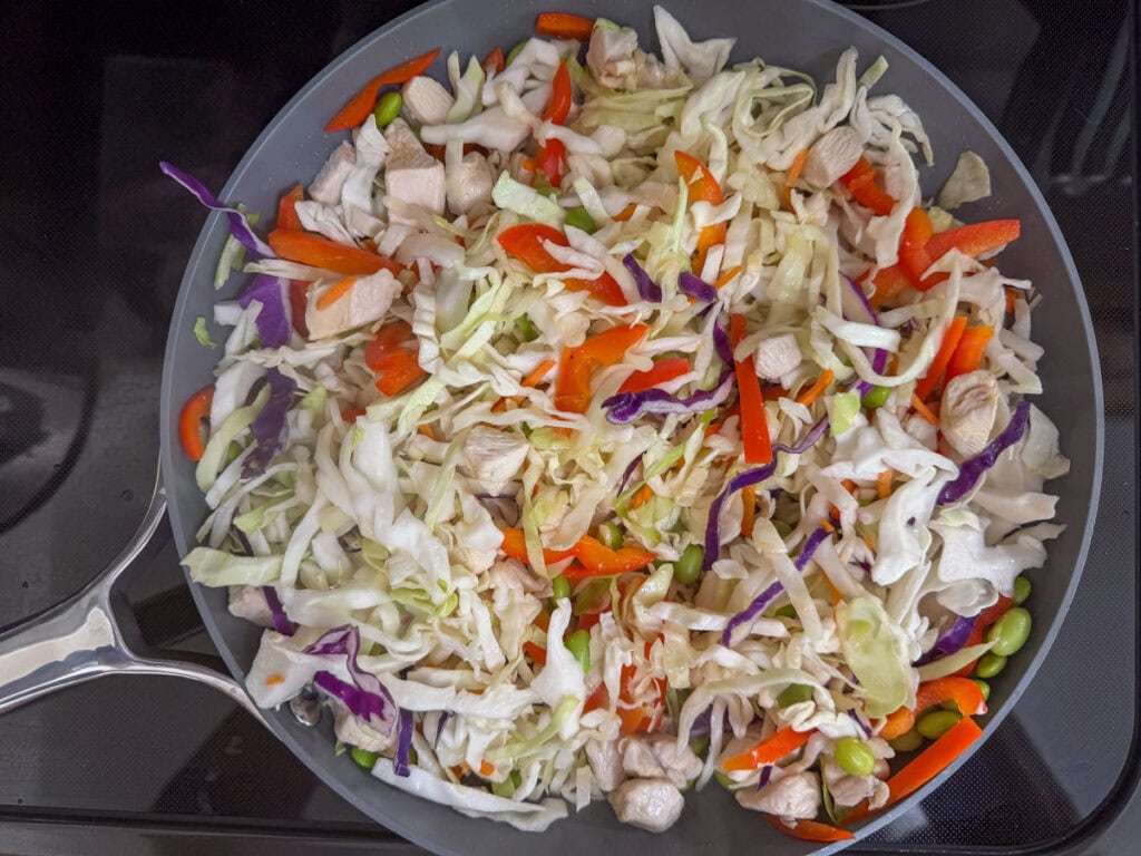 Shredded cabbage added to the chicken, bell peppers and edamame in the skillet before the cabbage has been cooked.