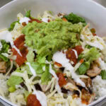 Closeup overhead shot of a Chipotle Chicken Burrito Bowl where the rice, chicken, black beans, shredded cheese, lettuce, salsa, guacamole and crema are visible.